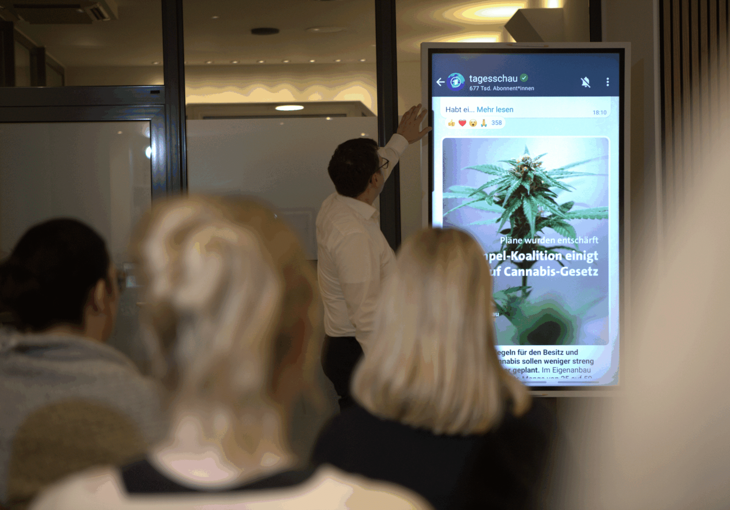 _MGL2469 „Mann präsentiert vor Publikum eine Nachrichtenmeldung zum neuen Cannabis-Gesetz auf einem großen Bildschirm.“